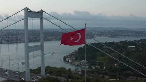 Turkish-Flag-Waving-in-Wind-in-front-of-Istanbul-Bosphorus-Bridge,-Aerial-medium-shot-slide-right